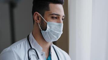 Male pensive thoughtful arab man therapist surgeon wear white uniform medical face mask standing at workplace looking out window preventing spread infection pandemic outbreak close-up doctor portrait photo