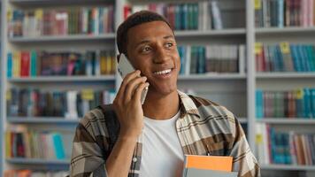 Happy African American man talking mobile phone smartphone technology communicate conversation library student learner university chatting smiling male guy bookshelf educational study calling laughter photo