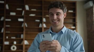 Happy wide smiling young Caucasian male business man businessman guy entrepreneur using mobile phone smartphone typing message chatting browsing internet enjoy online app game ordering delivery indoor photo