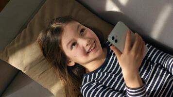 Caucasian European little girl lying on sofa holding using mobile phone smartphone child gadget online texting sms social networks smiling laughing enjoyment watching funny internet chatting kid photo