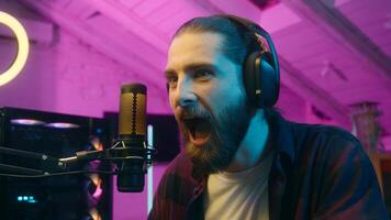 Focused glad Caucasian man gamer stream commenting game in microphone look monitor male play virtual strategy online cyber sport addict tense player blogger screaming win successful match victory home photo