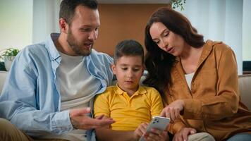 Happy family father mother parents son kid boy child talk communicate looking smartphone screen together on couch home using mobile app for play watch on Internet browsing social media on phone photo