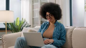 Young African American girl working distant from home with laptop at sofa ethnic woman freelancer typing email on computer online job using web service for e-learning study on couch user browsing net photo