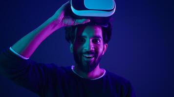 Arabian man take off VR glasses excited smile amazed with metaverse virtual reality cyberspace Indian guy with 3d goggles helmet happy wow reaction after play game meta universe neon ultraviolet space photo