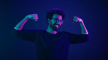 Funny Indian man slim guy fooling around show hands muscle biceps sport strong power bodybuilder ultraviolet neon studio background proud champion boast efficient security coding achievement level up photo
