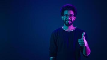 Smiling Indian man amazed Arabian IT guy Muslim male posing pointing index finger on copy space workspace isolated on neon dark blue ultraviolet background in studio showing thumb up coding promotion photo