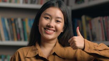Female portrait smiling happy asian woman girl student 20s looking at camera in university library showing thumbs up like gesture approval symbol successful feedback recommendation agree great result photo
