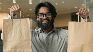 Happy joyful indian man with glasses indoors male buyer client guy customer shopper looking camera smiling showing paper bags satisfied purchases discount sale in store shows purchase goods packages photo