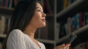 Asian woman female ethnic girl student searching book in university library on bookshelf using phone task list checking lesson schedule use mobile application on smartphone modern technology education photo