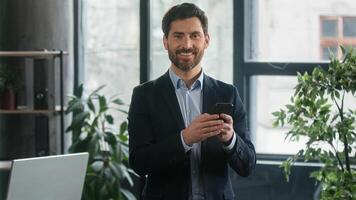 Caucasian man businessman business male using phone in office checking email browsing social network making order in online store choosing delivery chatting client on smartphone looking camera smiling photo
