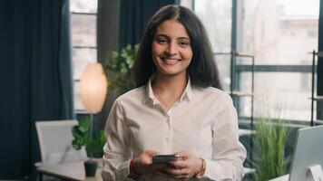 contento multirracial mujer utilizando móvil teléfono en oficina sonriente mirando a cámara mujer de negocios indio niña árabe hembra ejecutivo trabajador teléfono inteligente Internet surf hojeada en línea comprobación Noticias sonrisa foto