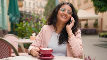 contento sonriente indio árabe étnico mujer niña visitante Bebiendo taza café en ciudad café Mañana al aire libre hembra mujer de negocios hablando móvil teléfono negocio llamada simpático hablar teléfono inteligente discutir Noticias foto