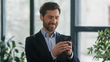 Caucasian man businessman business male using phone in office checking email browsing social network making order in online store choosing delivery chatting client on smartphone looking camera smiling photo