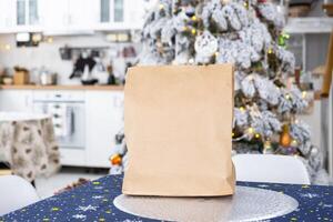 A kraft paper bag is on the kitchen table. Delivery of sushi and rolls, cookies, gift in the festive interior of the house with a white Christmas tree for Christmas and New year. photo