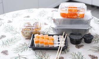 Delivery of sushi and rolls in food containers on the table in the festive interior of the kitchen of the house with a white Christmas tree for Christmas and New year. Food delivery for the holidays photo