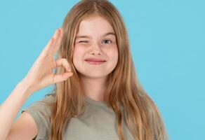 Funny And joyful child girl shows okay sign and winks looking at camera isolated on blue background photo