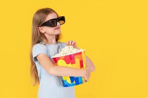 Kids cinema. little girl with popcorn in 3d glasses looking at camera and wondering on yellow background, kid amazed viewer watching movie photo