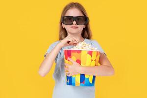 Kids cinema. little girl with popcorn in 3d glasses looking at camera and wondering on yellow background, kid amazed viewer watching movie photo