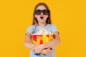 Kids cinema. Shocked little girl with popcorn in 3d glasses looking at camera and wondering on yellow background, kid amazed viewer watching movie photo