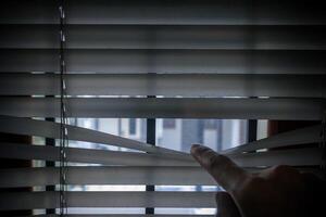 Peeking through window blinds. Hand separating the slats of the window curtain with fingers to look out. photo