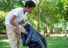 A boy of dedicated volunteers are participating in an environmental cleaning activity in a park. Boy wearing gloves and hold large black bags. Boy collecting trash and plastic bottles in a park. photo