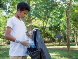 A boy of dedicated volunteers are participating in an environmental cleaning activity in a park. Boy wearing gloves and hold large black bags. Boy collecting trash and plastic bottles in a park. photo