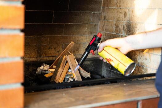 This image shows a fireplace being lit using a butane torch. The flame from the torch is helping ignite pieces of wood arranged in the fireplace. photo