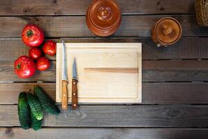 Top-down view, variety of fresh tomatoes and cucumbers next to a wooden cutting board with two knives. Clay pot, adding to the rustic vibe. photo