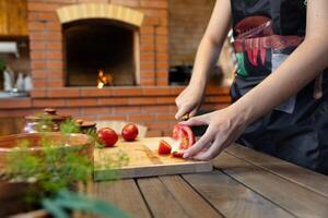 Cutting tomatoes on a wooden cutting board. Cozy kitchen with a brick oven or fireplace in the background, adding warmth. Fresh herbs in the foreground. photo