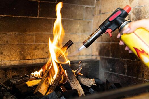 This image shows a fireplace being lit using a butane torch. The flame from the torch is helping ignite pieces of wood arranged in the fireplace. photo