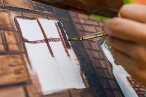 Close-up of someone skillfully painting on a canvas. The artwork looks like it depicts a rustic scene, possibly featuring elements like bricks or wood, giving it a cozy, homely feel. photo