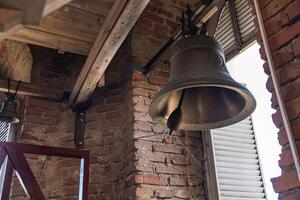 Large metal bell hanging inside a structure with brick walls and wooden beams. The architecture has a rustic feel, reminiscent of traditional or historical buildings. photo