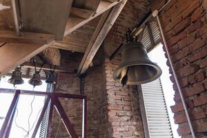 Large metal bell hanging inside a structure with brick walls and wooden beams. The architecture has a rustic feel, reminiscent of traditional or historical buildings. photo