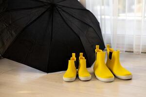 Bright yellow rubber boots for children and adults placed under a black umbrella indoors photo