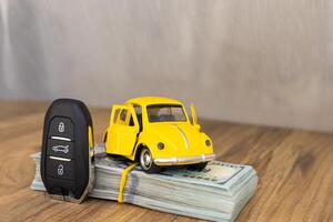 A small yellow toy car on top of a stack of cash with a car key beside it in a well-lit setting photo