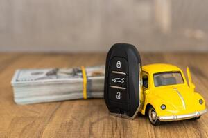 A small yellow toy car on top of a stack of cash with a car key beside it in a well-lit setting photo