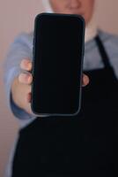 Woman in apron holds blank smartphone while standing in softly lit room photo