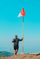 An adventurer is at the top of the ancient volcano Nglanggeran which is dominated by rocks with the Indonesian national flag. Photo of adventurer reaching rocky mountain peak with flag waving