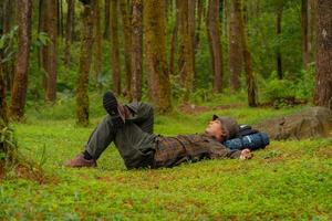 Asian adventurers enjoy the serenity of the wild by resting on the green grass in the pine forest area. A male adventurer was lying on the green grass in a quiet pine forest area. photo