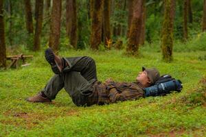Asian adventurers enjoy the serenity of the wild by resting on the green grass in the pine forest area. A male adventurer was lying on the green grass in a quiet pine forest area. photo