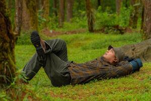 Asian adventurers enjoy the serenity of the wild by resting on the green grass in the pine forest area. A male adventurer was lying on the green grass in a quiet pine forest area. photo