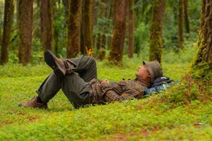 Asian adventurers enjoy the serenity of the wild by resting on the green grass in the pine forest area. A male adventurer was lying on the green grass in a quiet pine forest area. photo