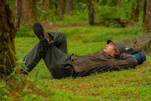 Asian adventurers enjoy the serenity of the wild by resting on the green grass in the pine forest area. A male adventurer was lying on the green grass in a quiet pine forest area. photo