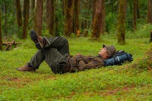 Asian adventurers enjoy the serenity of the wild by resting on the green grass in the pine forest area. A male adventurer was lying on the green grass in a quiet pine forest area. photo