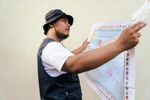 Half body portrait of a male model posing like an adventurer while looking at a map. Asian adult man wearing casual clothes with hat and vest is looking at a sheet of world map on a beige background. photo