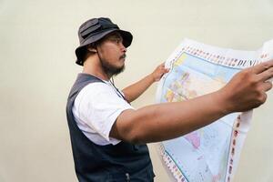 Half body portrait of a male model posing like an adventurer while looking at a map. Asian adult man wearing casual clothes with hat and vest is looking at a sheet of world map on a beige background. photo