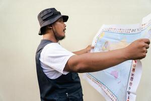 Half body portrait of a male model posing like an adventurer while looking at a map. Asian adult man wearing casual clothes with hat and vest is looking at a sheet of world map on a beige background. photo