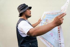 Half body portrait of a male model posing like an adventurer while looking at a map. Asian adult man wearing casual clothes with hat and vest is looking at a sheet of world map on a beige background. photo