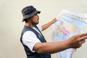 Half body portrait of a male model posing like an adventurer while looking at a map. Asian adult man wearing casual clothes with hat and vest is looking at a sheet of world map on a beige background. photo