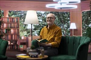 Elderly man reading a book sitting alone in his lounge area at home, relaxing and feeling good with free time. Person with an intellectual hobby, passionate about literature and lecture. photo